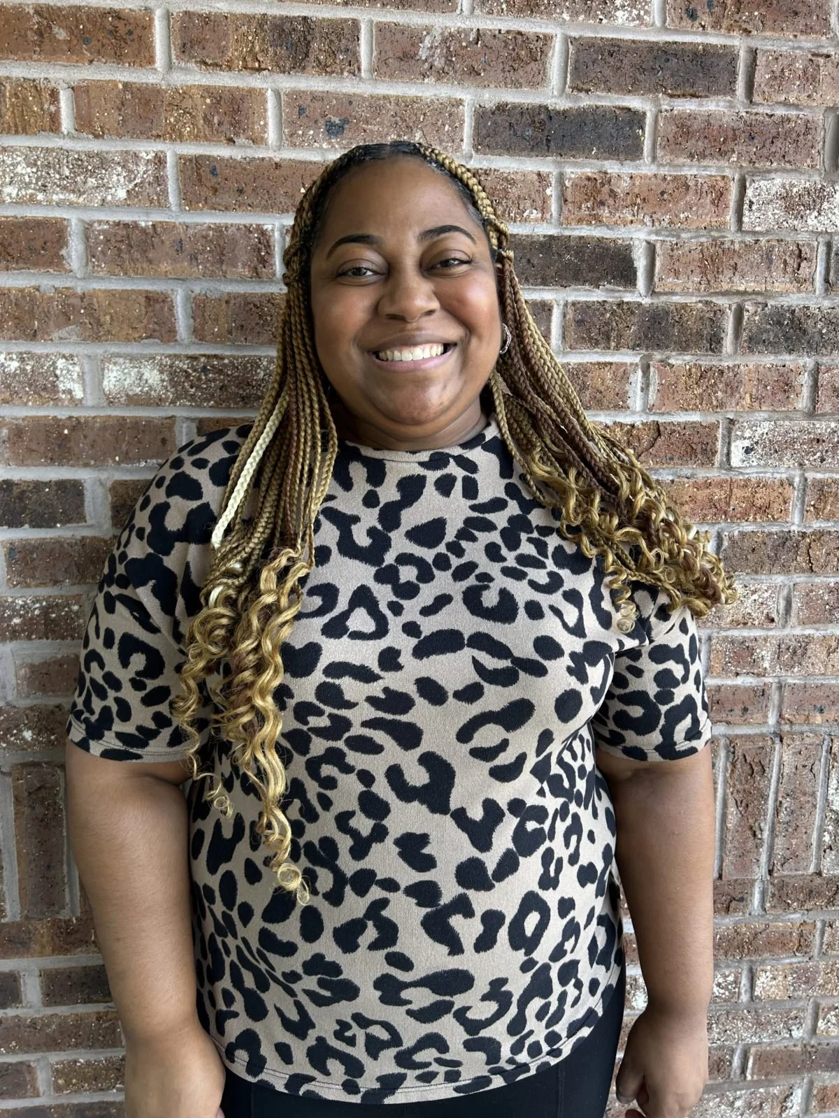 A woman with long blonde styled braids smiling in front of a brick wall, wearing a beige and black leopard print shirt.