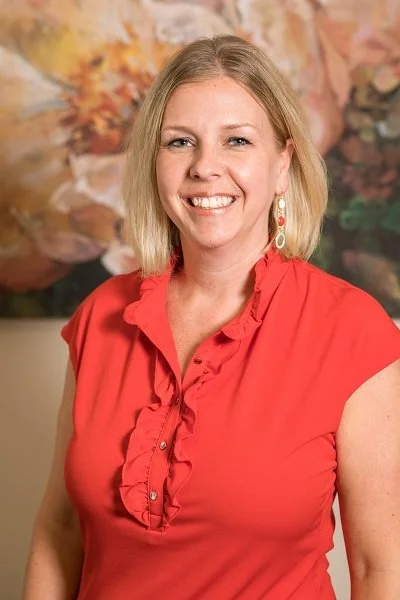 A woman with shoulder-length blonde hair smiling, wearing a coral red blouse with ruffles and dangling earrings, standing in front of a blurred colorful floral background.