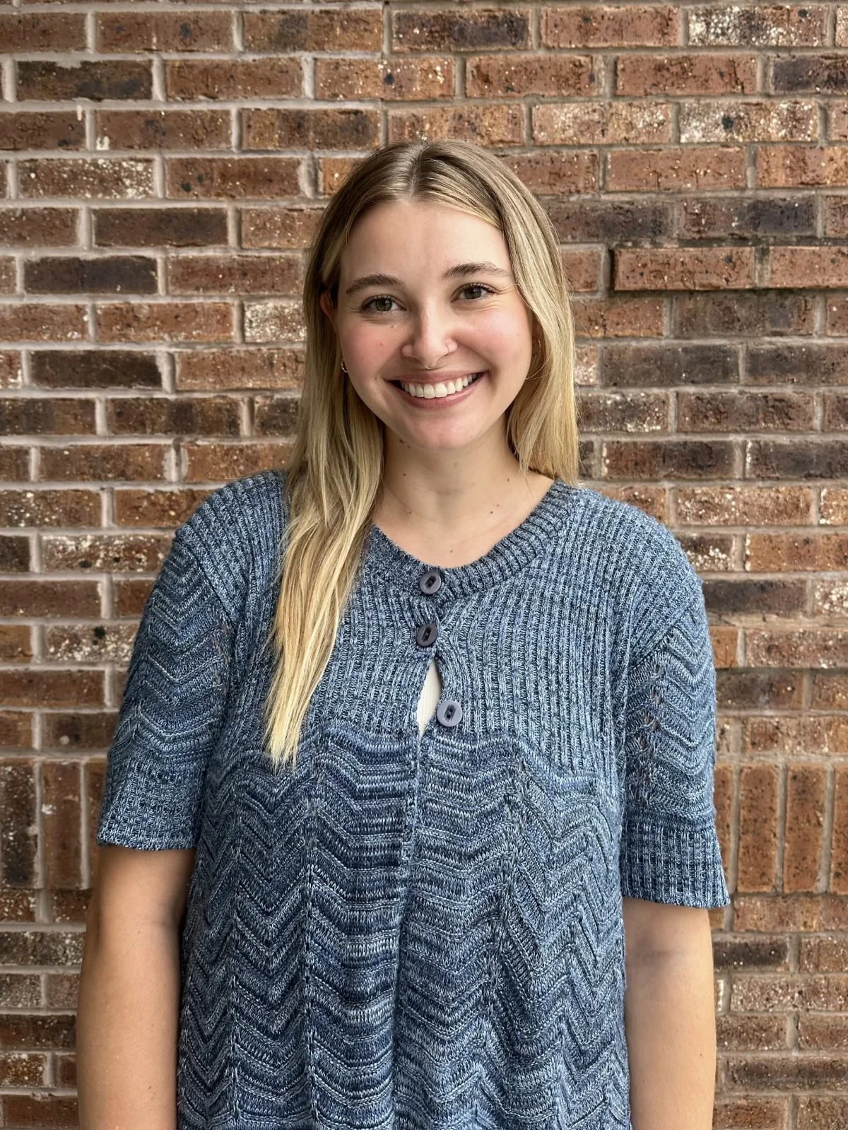 A young woman with blonde hair smiling, wearing a blue patterned cardigan, standing against a brick wall.