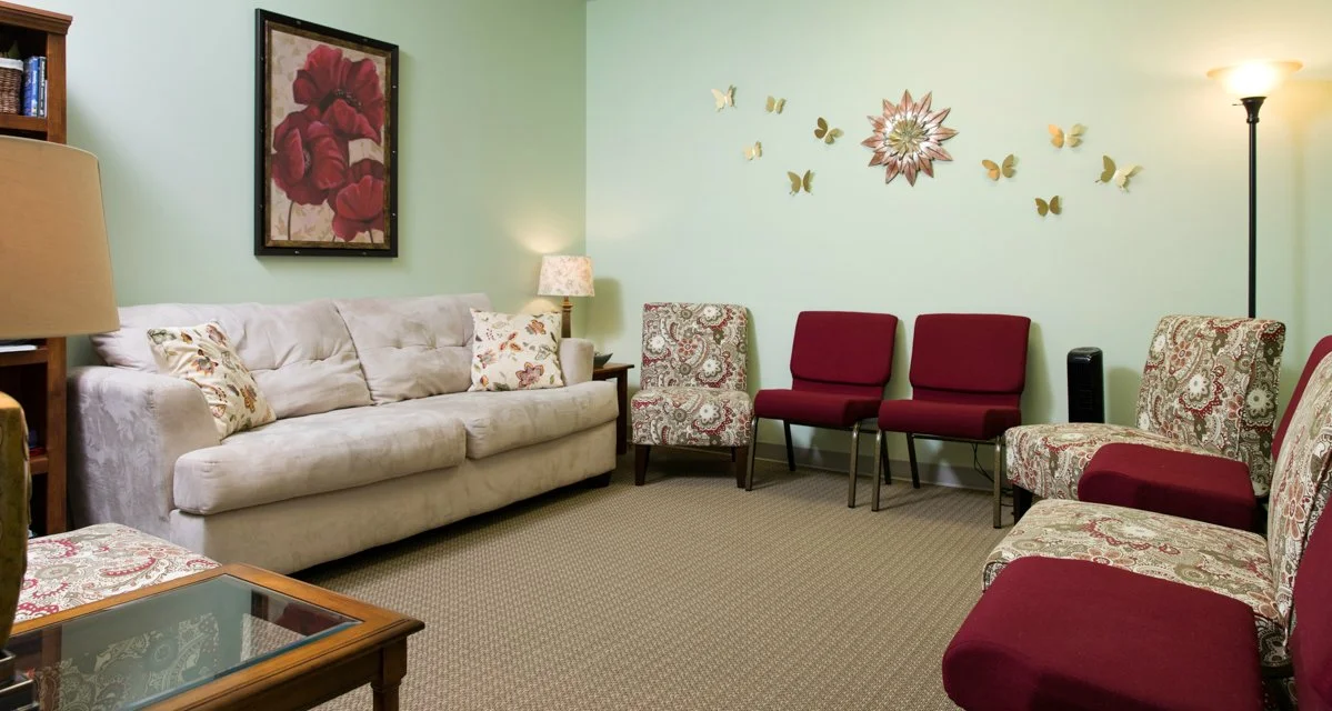Living room with a beige sofa, patterned armchairs, red and patterned chairs, a framed floral painting, a wall mirror, a floor lamp, a side table with a lamp, a standing lamp, and butterfly wall decorations.