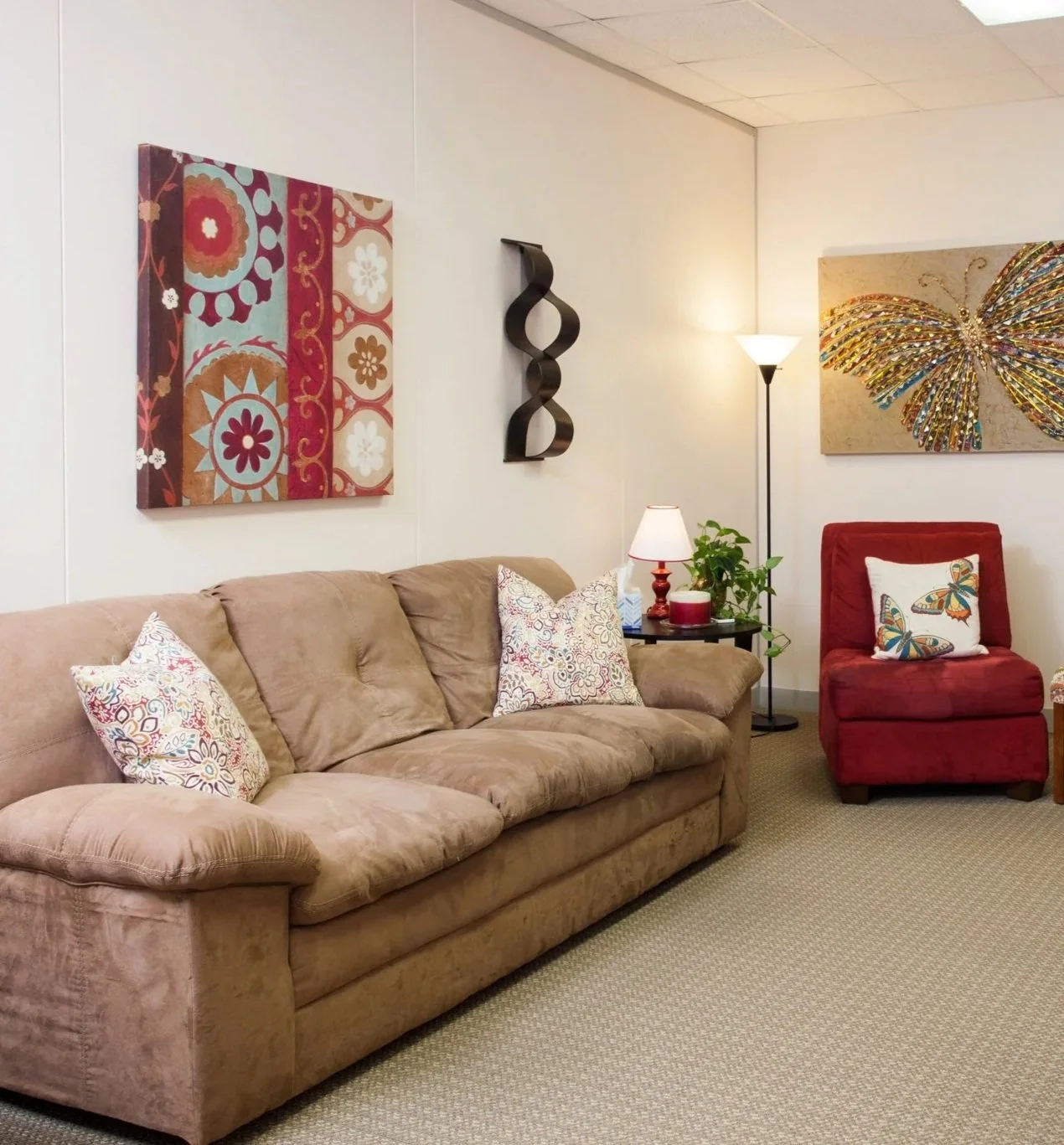 Living room with a beige sofa with three decorative pillows, a small side table with a lamp and plants, a red armchair with a butterfly pillow, wall art including a colorful butterfly, floral painting, and abstract sculpture, and a standing lamp in the corner.