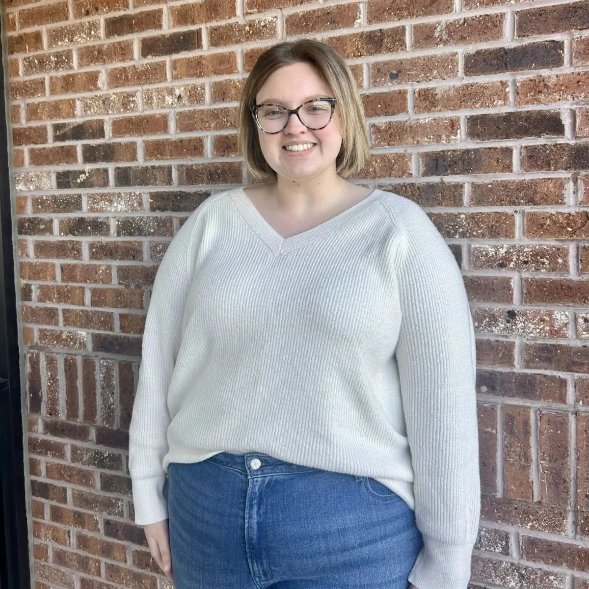 A young woman with short light brown hair, glasses, wearing a white sweater and blue jeans, standing against a brick wall, smiling.