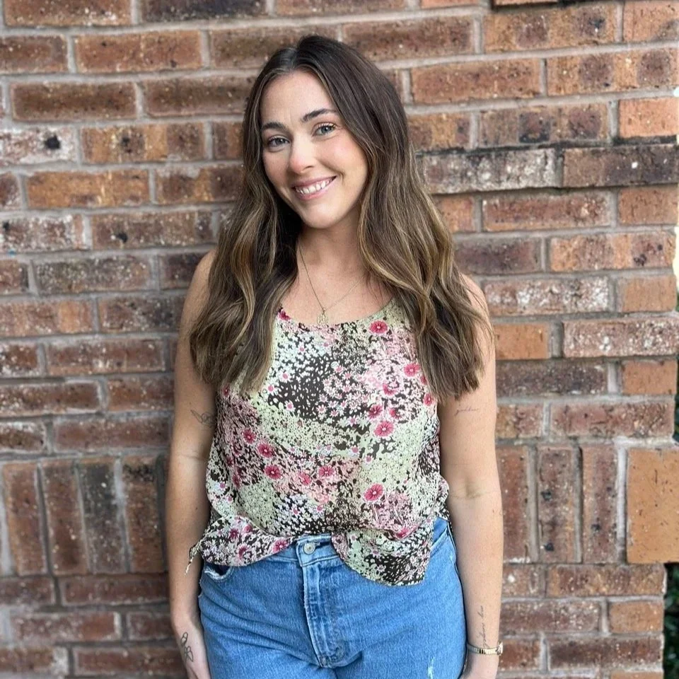 A young woman with long brown hair, smiling, standing against a brick wall. She is wearing a floral sleeveless top and blue jeans.