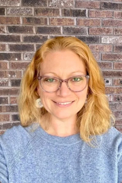 A woman with shoulder-length blonde hair smiling, wearing a red top and a long necklace with a teardrop pendant, standing outdoors in front of a weathered, greenish wall.