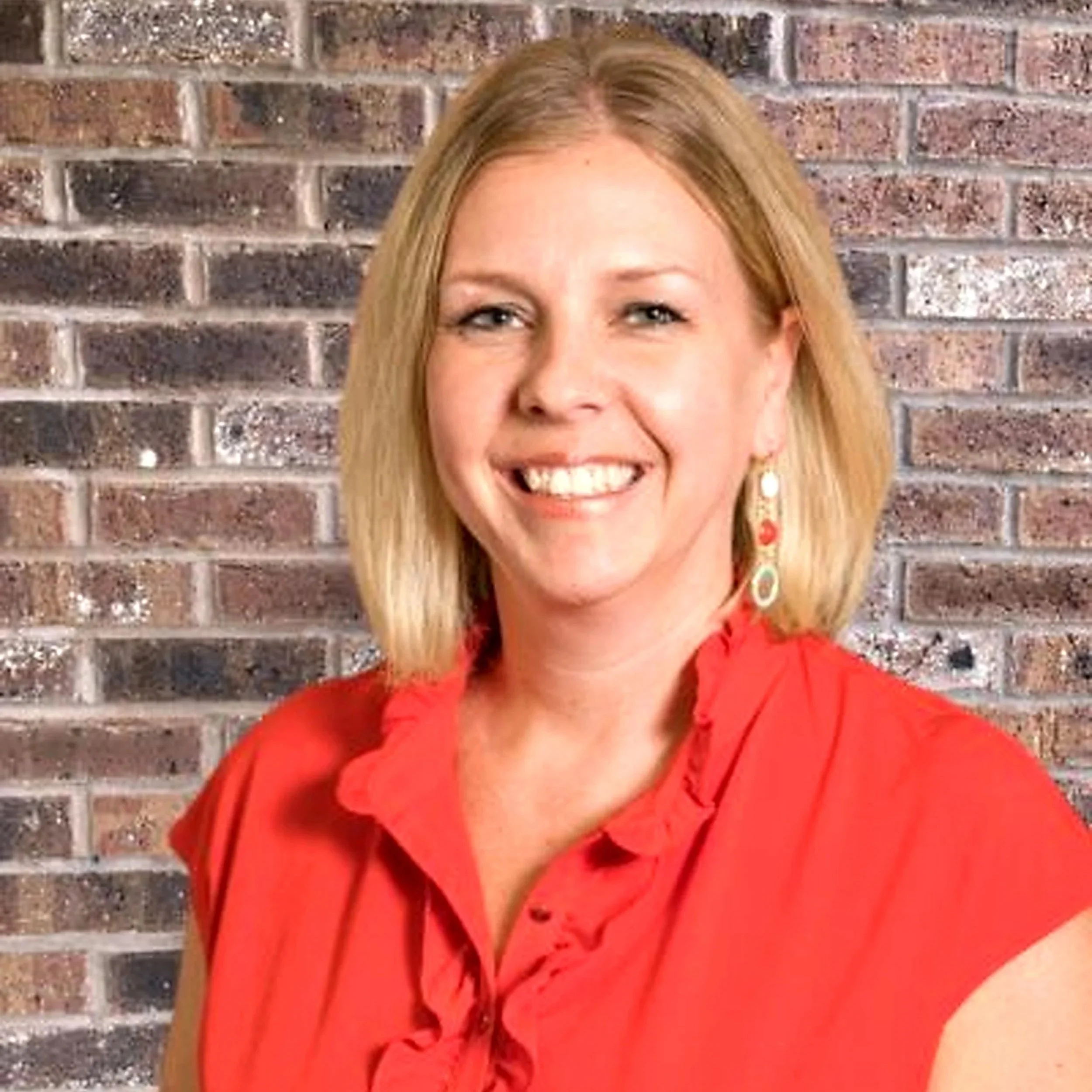 A woman with blond hair smiling, wearing a red top, standing in front of a colorful floral background.