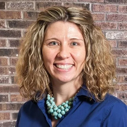 A woman with curly blonde hair smiling, wearing a blue shirt and a turquoise beaded necklace, with a colorful, abstract background.
