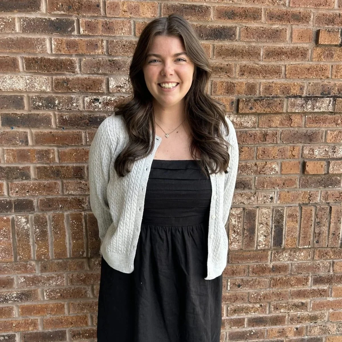 A young woman with long brown hair smiling, standing in front of a brick wall, wearing a black dress, a white cardigan, and a delicate necklace.