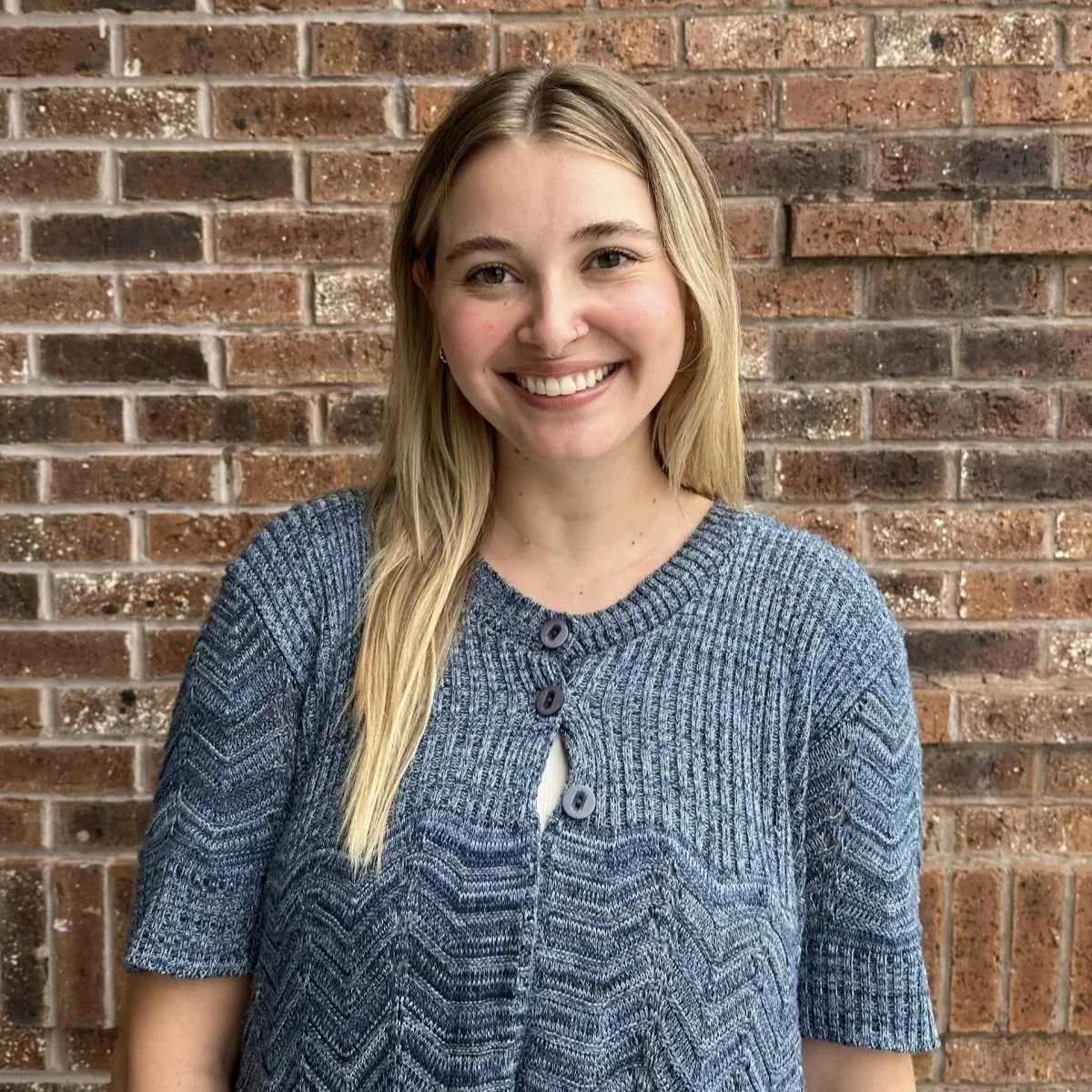 A young woman with long blonde hair, smiling, in front of a brick wall, wearing a blue knitted top.