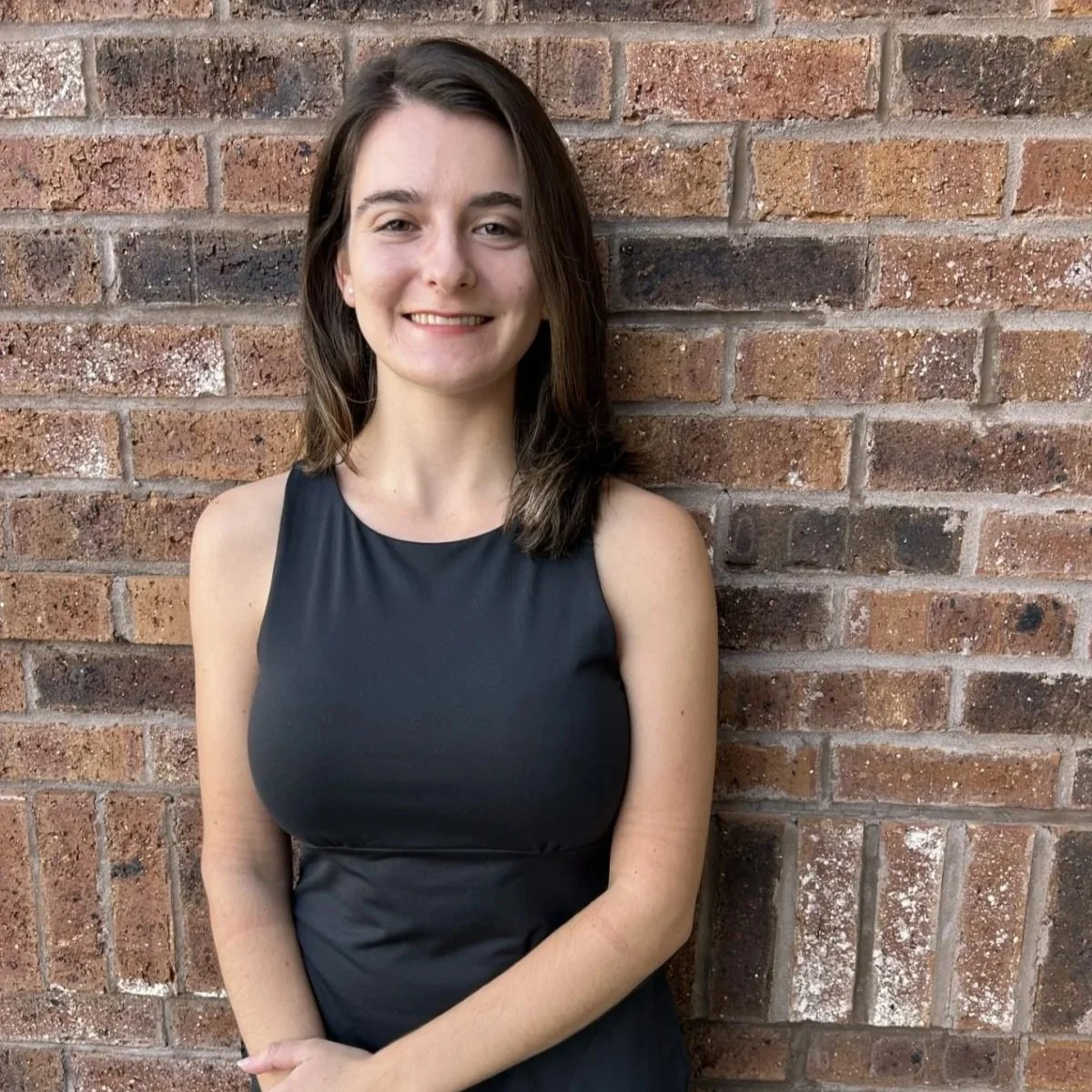 Young woman with shoulder-length brown hair smiling, wearing a sleeveless black dress, standing in front of a brick wall.