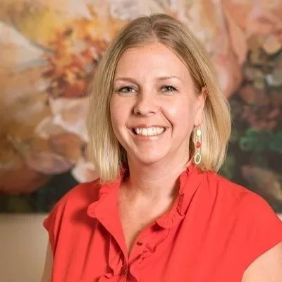A smiling woman with shoulder-length blonde hair, wearing a red blouse and earrings, standing in front of a colorful floral background.