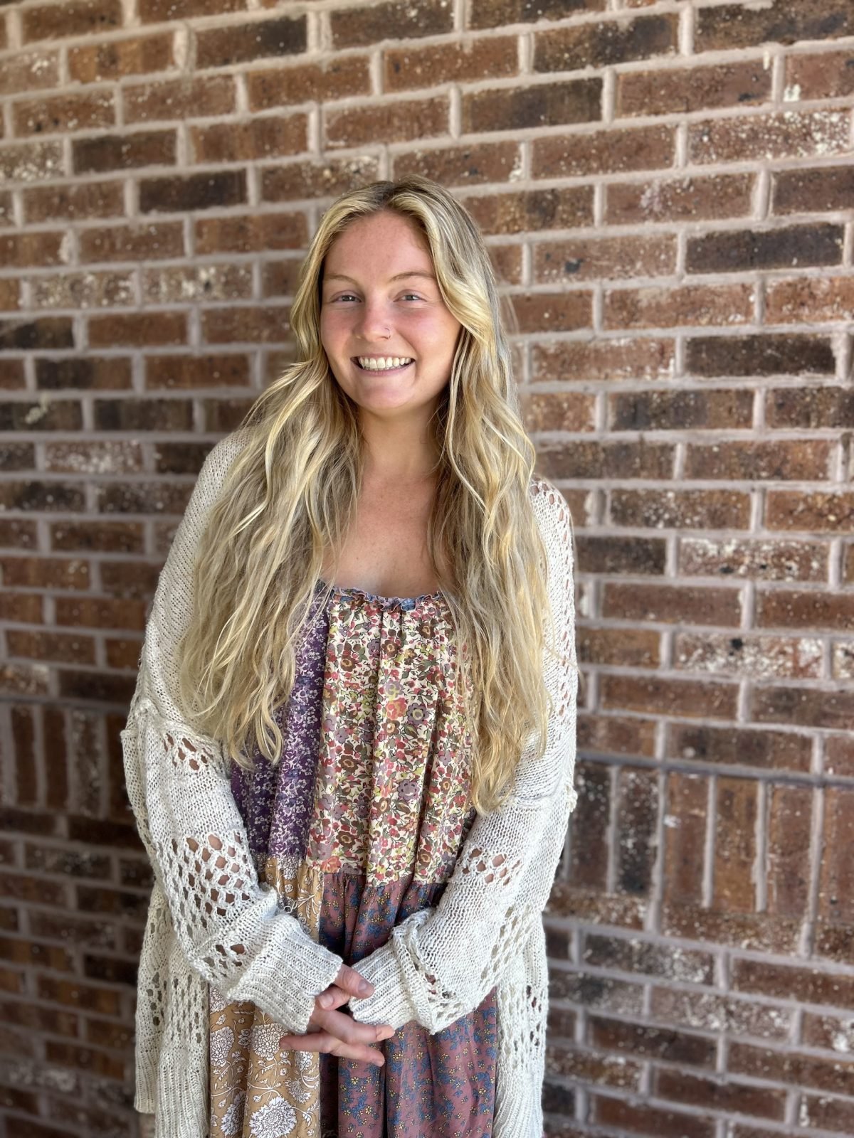 A smiling woman with long wavy blonde hair standing in front of a brick wall, wearing a patterned dress and an open knit cardigan.
