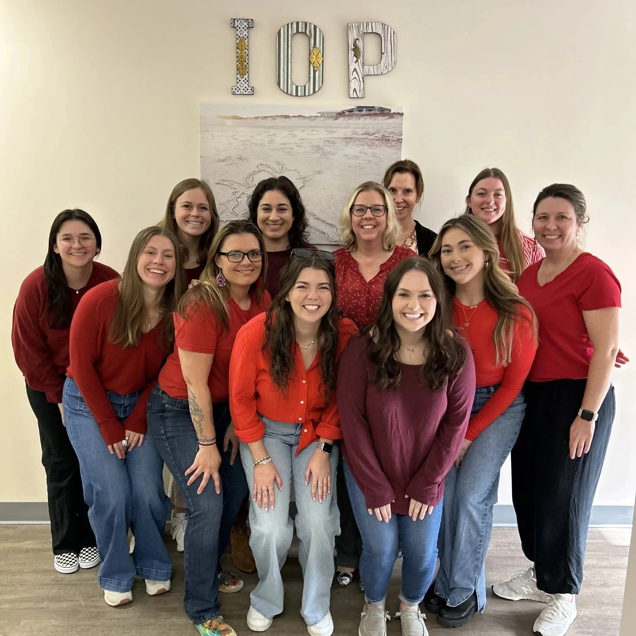 Group of twelve women smiling for a photo, standing in front of a wall with beach-themed artwork and colorful letters spelling 'TOP'.
