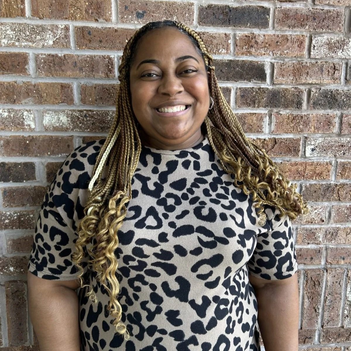 A woman smiling in front of a brick wall, wearing a leopard print top and having long blonde braided hair.