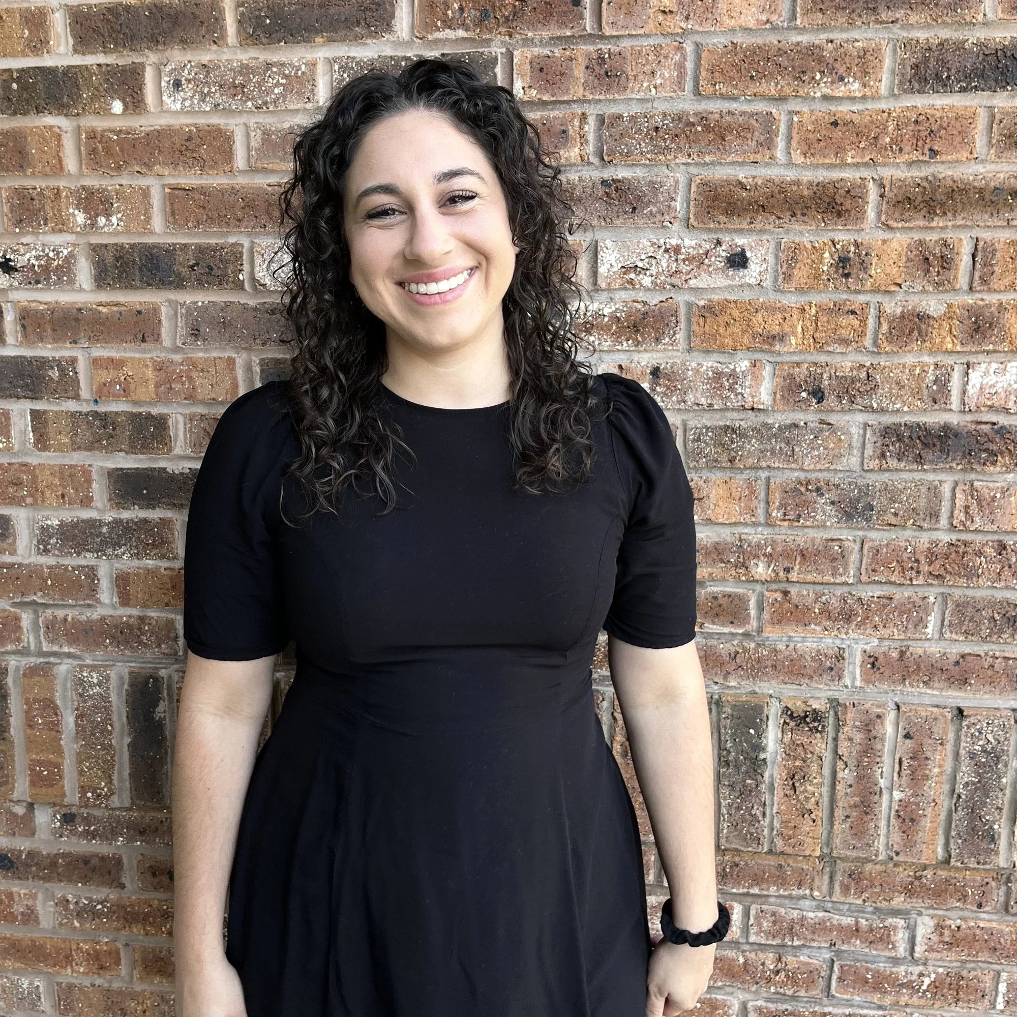 A woman with curly dark hair wearing a black dress, smiling in front of a brick wall.