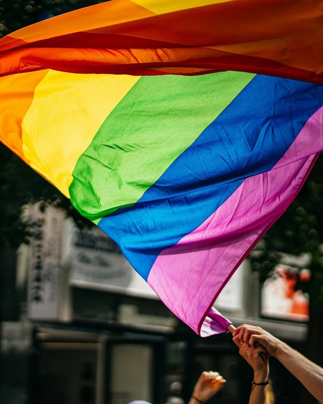 A rainbow pride flag being held up in a crowd during a Pride event, with a blurred urban background.