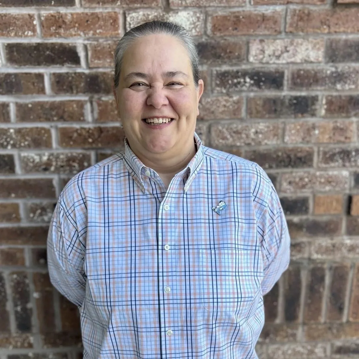 A middle-aged man smiling, wearing a plaid button-up shirt with a small airplane pin, standing in front of a brick wall.