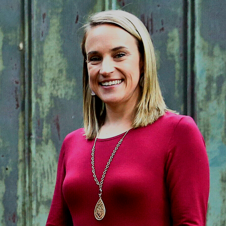 A woman with shoulder-length blonde hair smiling, wearing a red top and a long necklace with a teardrop pendant, standing outdoors in front of a weathered, greenish wall.