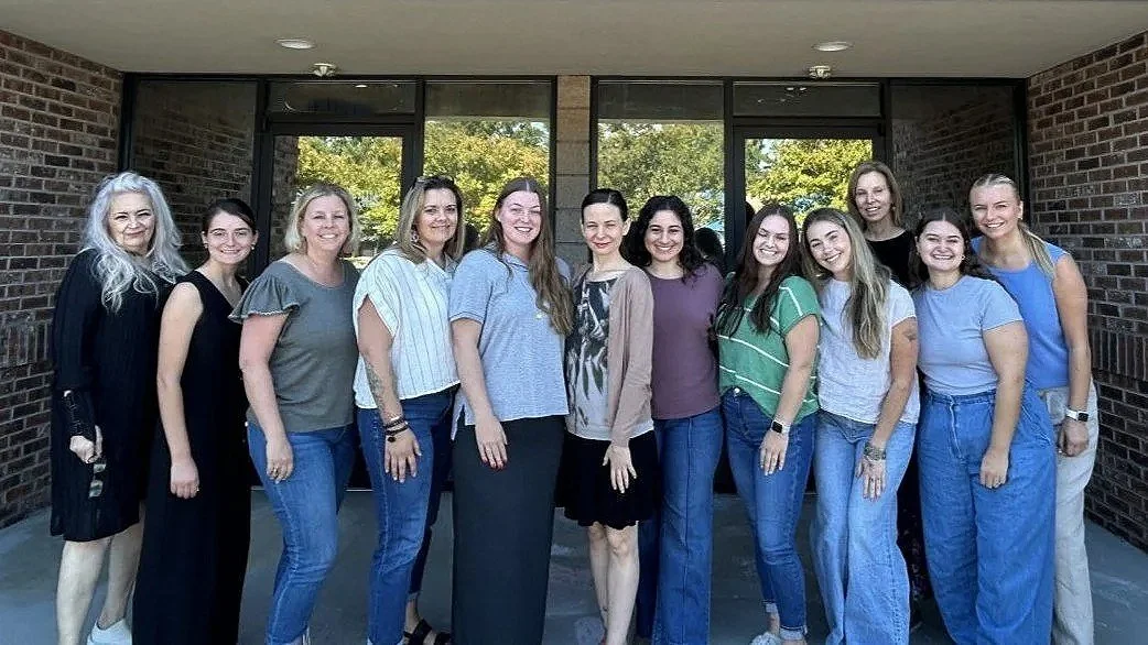 Group of fourteen women standing outdoors in front of a building with large windows, smiling at the camera.