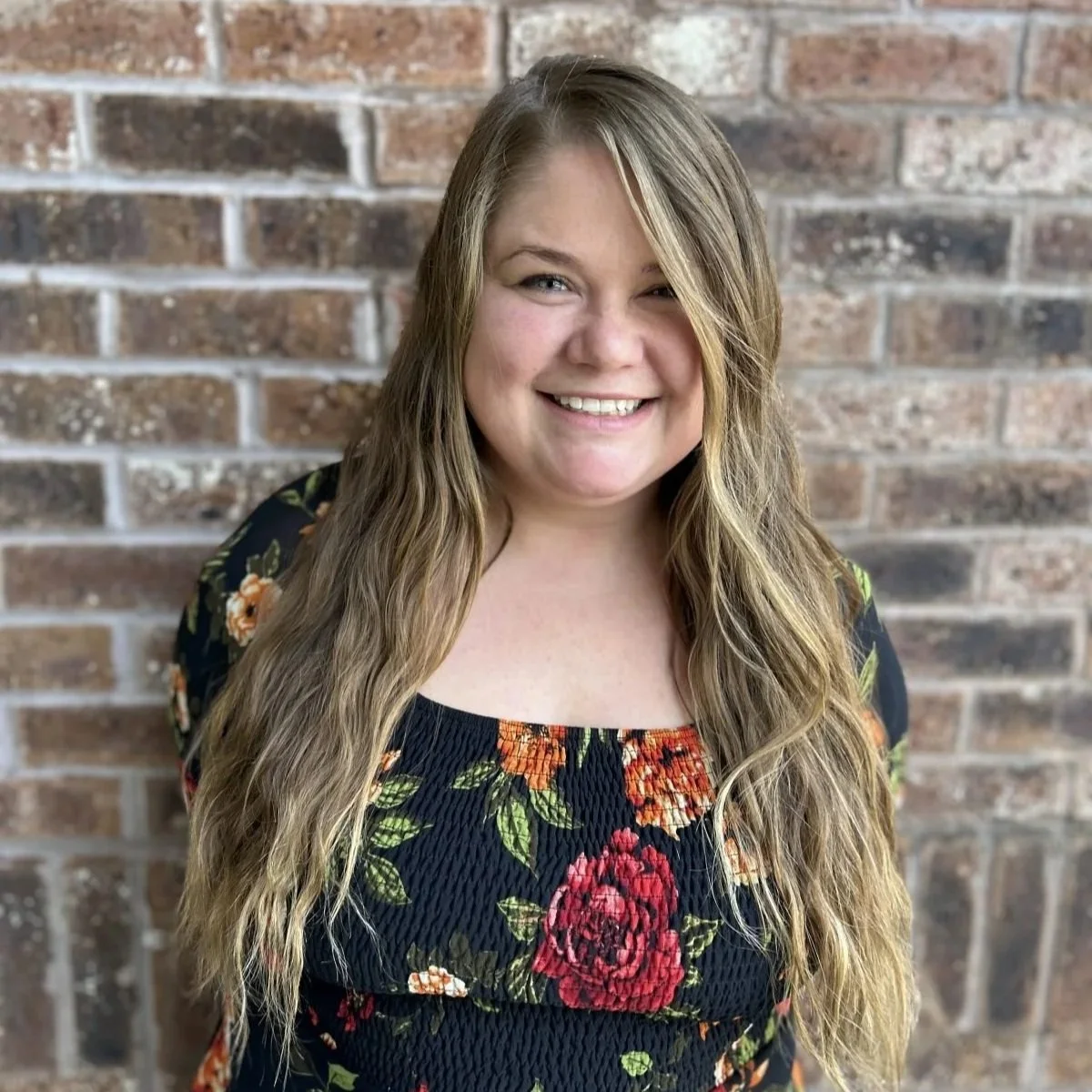 A young woman with long wavy blonde hair, smiling, standing in front of a brick wall, wearing a black floral blouse with orange, red, and green flowers.