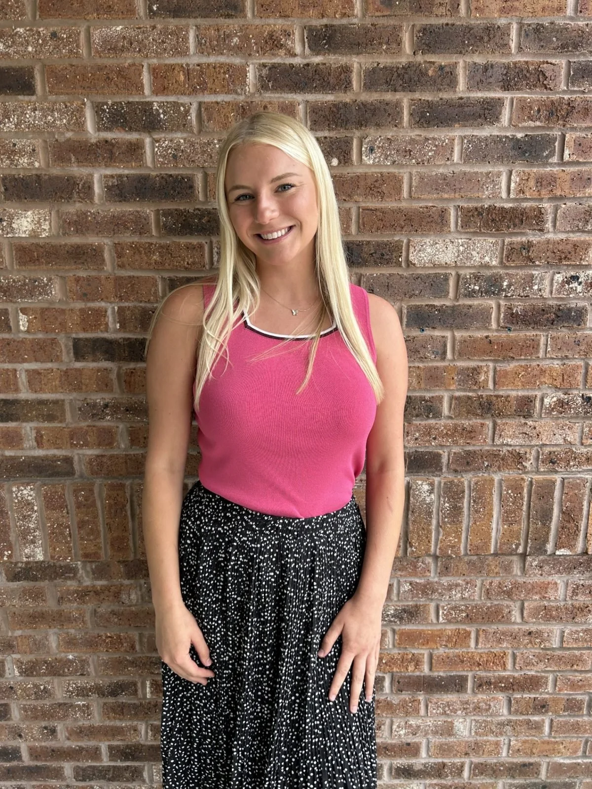A young woman with long blonde hair, wearing a pink sleeveless top and a black and white dotted skirt, standing against a brick wall and smiling at the camera.