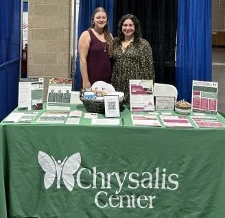Two women stand behind a table with informational pamphlets and materials at the Chrysalis Center, in front of blue curtains.