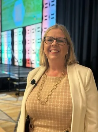 Smiling woman with glasses wearing a beige checkered top and white jacket, standing in front of a colorful digital display or screen in an indoor setting.