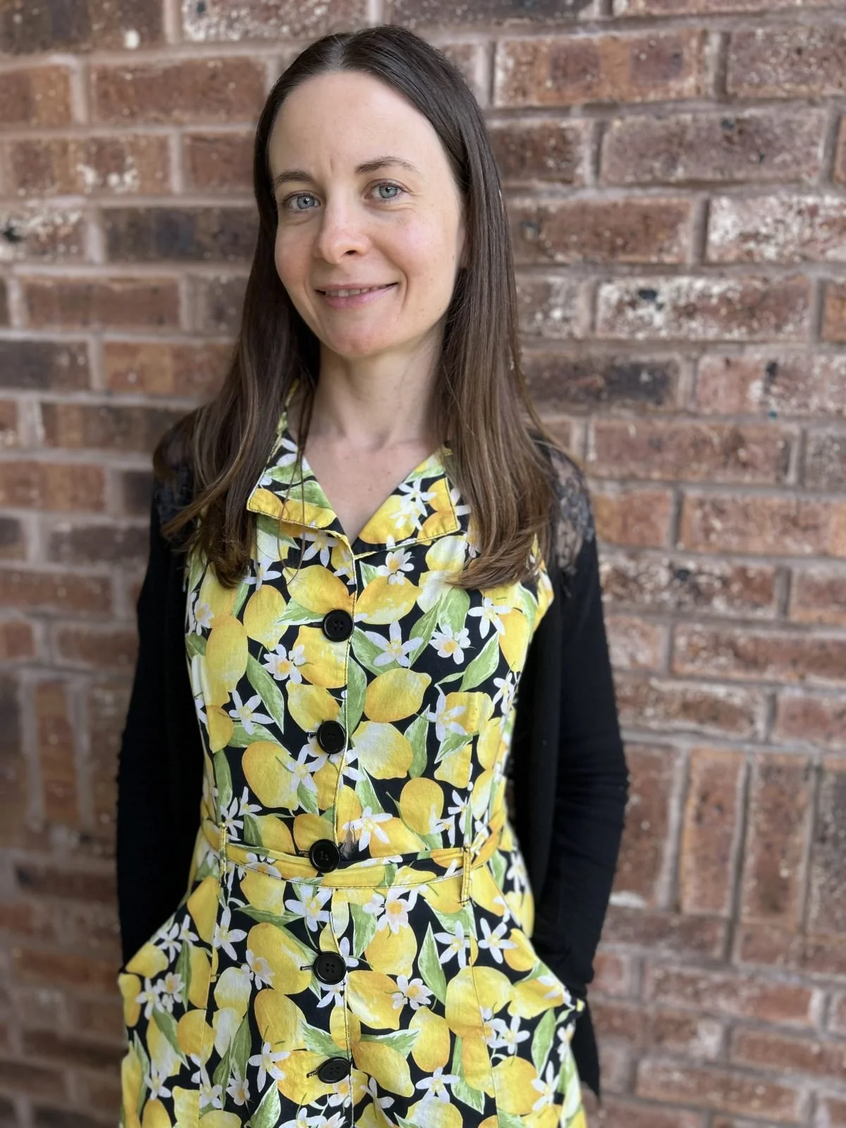 A woman with long brown hair and blue eyes stands in front of a brick wall, wearing a yellow, green, and white floral dress with black buttons and a black cardigan.
