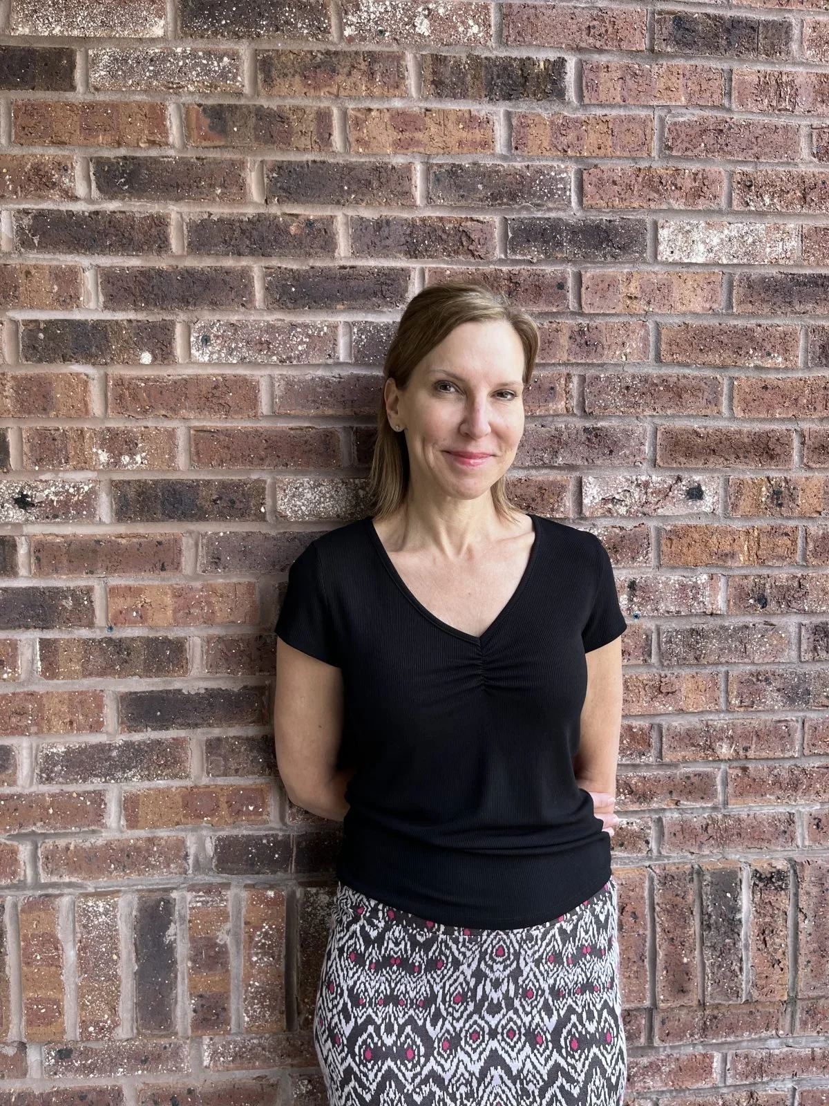 A woman with shoulder-length brown hair, wearing a black short-sleeve top and a patterned skirt, standing against a brick wall, smiling softly.