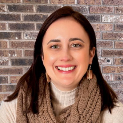A woman with dark brown hair smiling, wearing a beige sweater and chunky knit scarf, earrings, and in front of a colorful butterfly display.