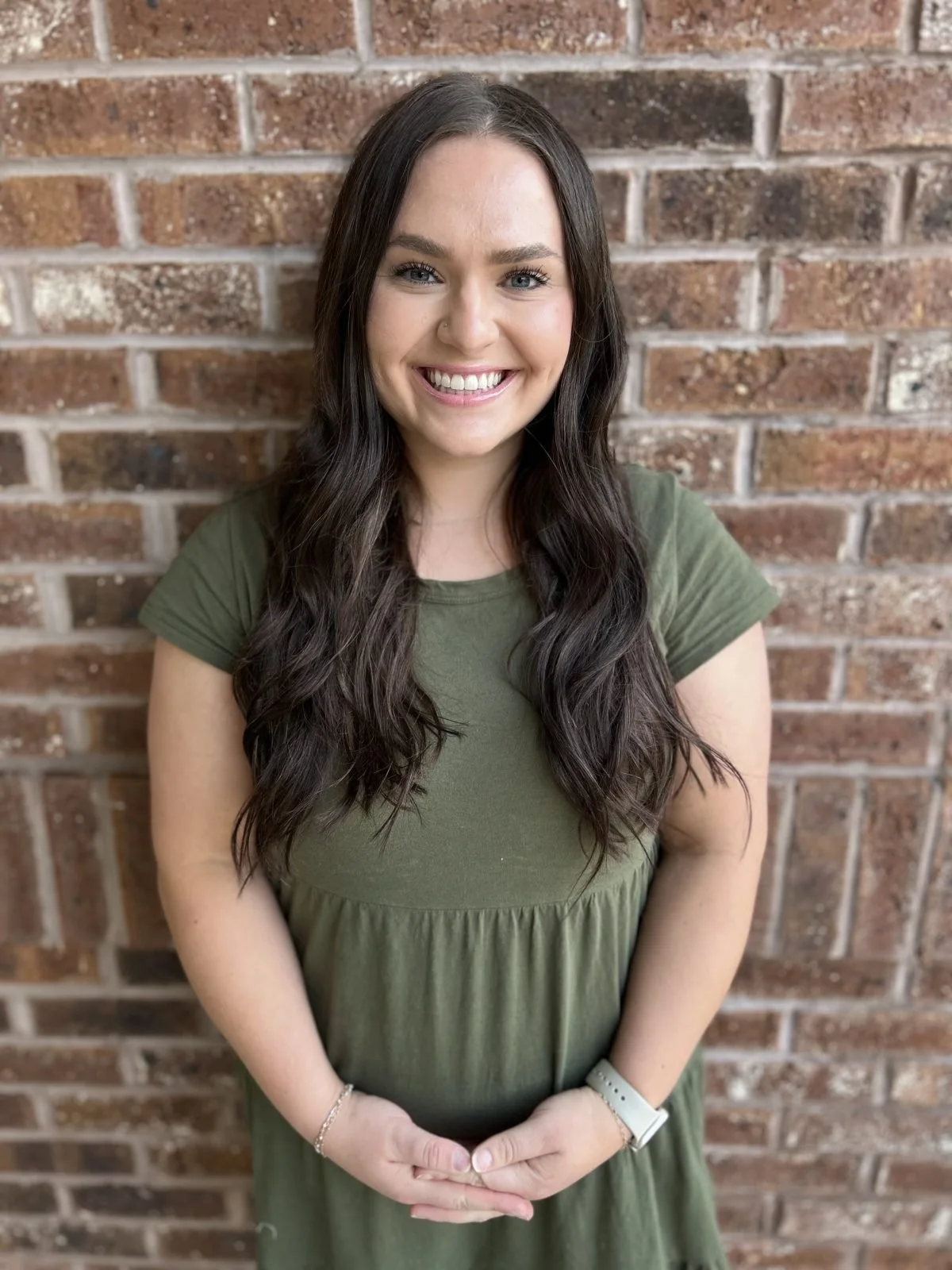 A smiling young woman with long, wavy dark hair, wearing a green dress and a white watch, standing in front of a brick wall.