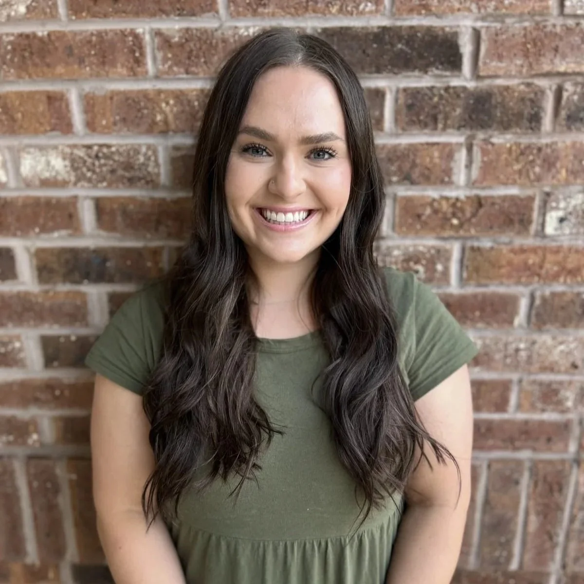 A young woman with long dark hair, smiling, wearing a green top, standing in front of a brick wall.