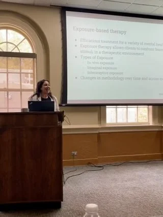 A woman stands at a podium giving a presentation in a room with large windows. A slide about exposure-based therapy is projected on a screen behind her.
