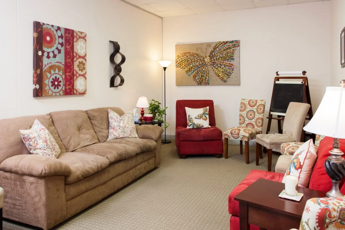 Living room with a beige couch, colorful wall art, a red armchair with a patterned pillow, side chairs with patterned upholstery, lamps, and an easel.
