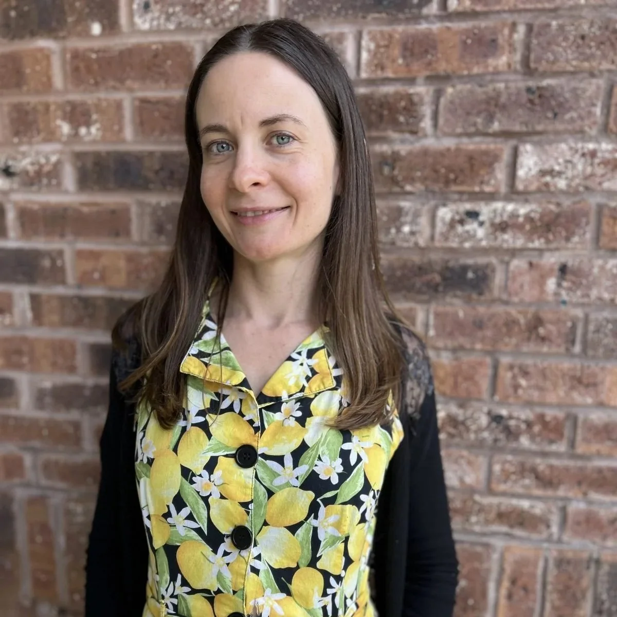 A woman with long brown hair and blue eyes standing in front of a brick wall, wearing a yellow lemon-patterned top with white flowers and a black cardigan.
