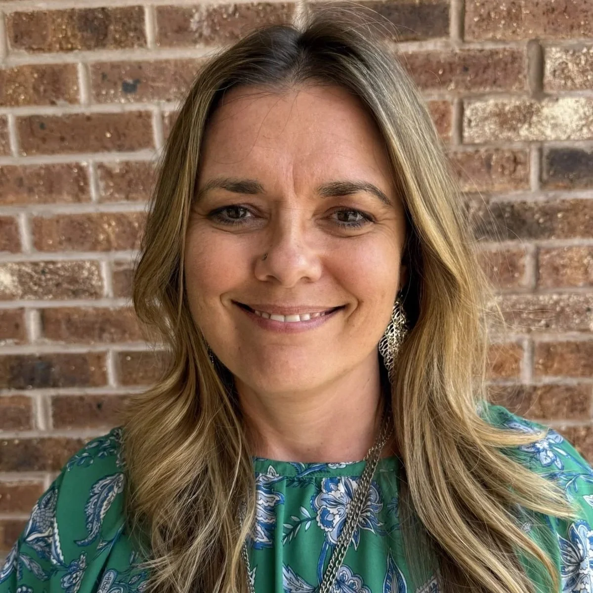 A woman with light brown, wavy hair and light skin, smiling, wearing a green dress with a blue and white floral pattern, earrings, and a necklace, standing in front of a brick wall.
