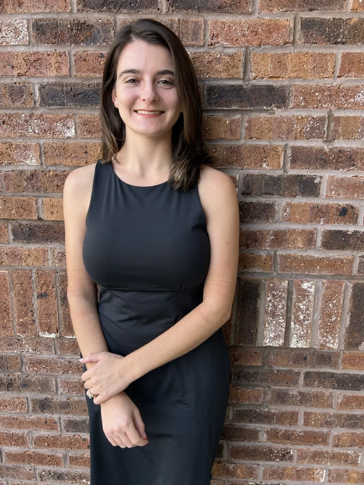 A young woman with shoulder-length brown hair, wearing a sleeveless black dress, standing against a brick wall, smiling at the camera.