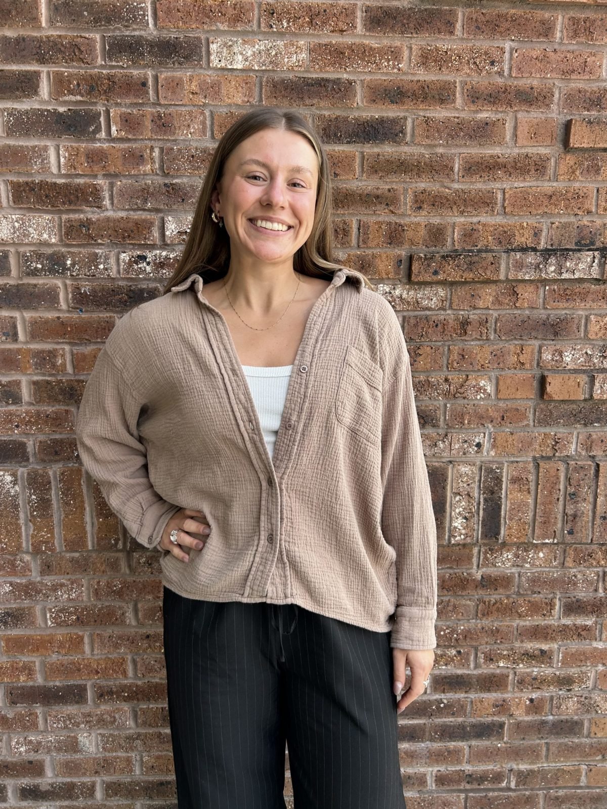 A woman with long brown hair smiling and standing against a red brick wall. She is wearing a beige button-up shirt over a white top and black pinstriped pants.