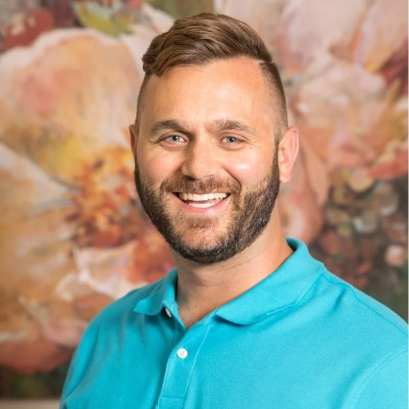 A smiling man with a beard and short brown hair, wearing a turquoise polo shirt, standing in front of an abstract, colorful painting.