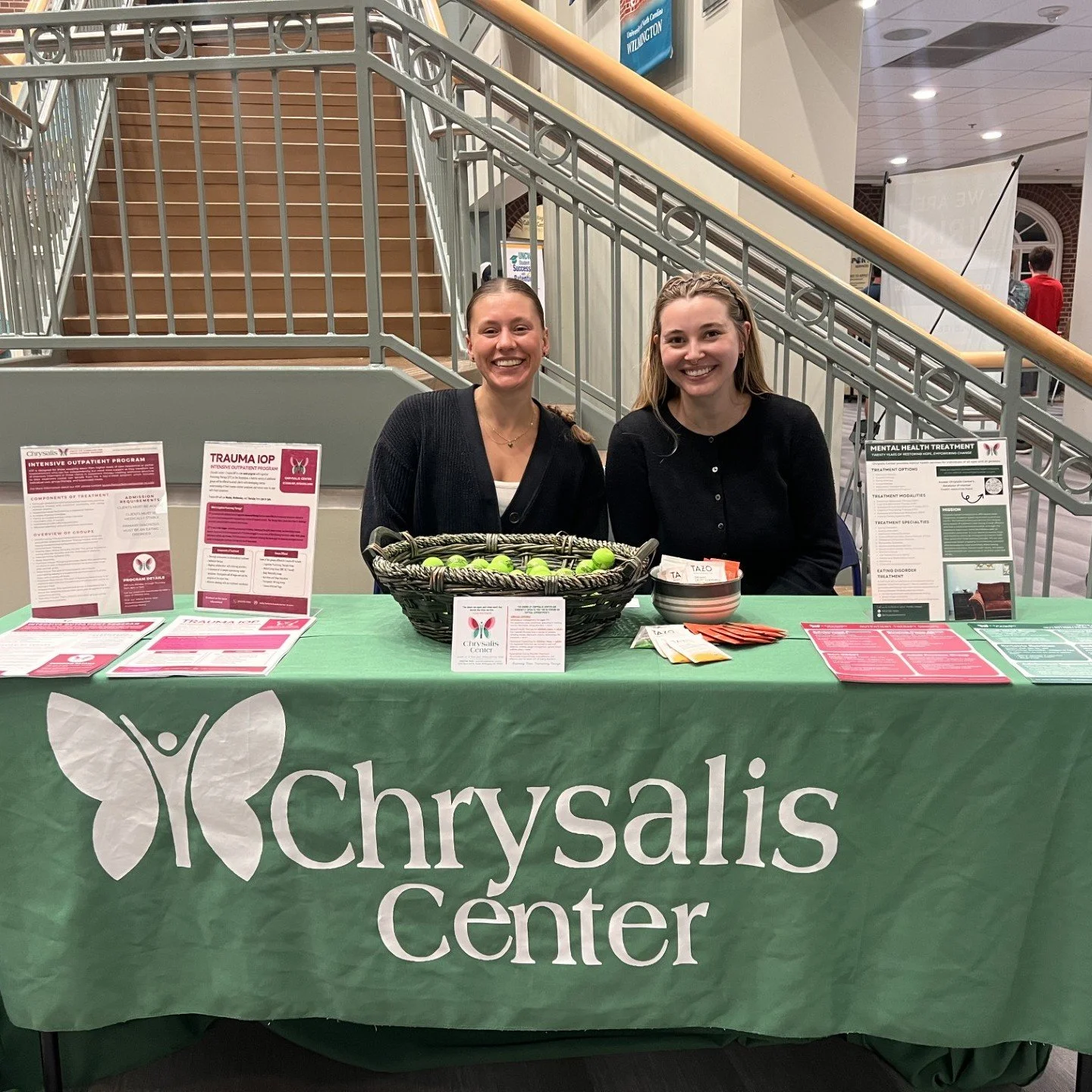 Two smiling women sitting behind a table with a green tablecloth that says 'Chrysalis Center.' The table has pamphlets, a basket of tennis balls, and a bowl of small items. They are in an indoor setting with stairs and a railing in the background.