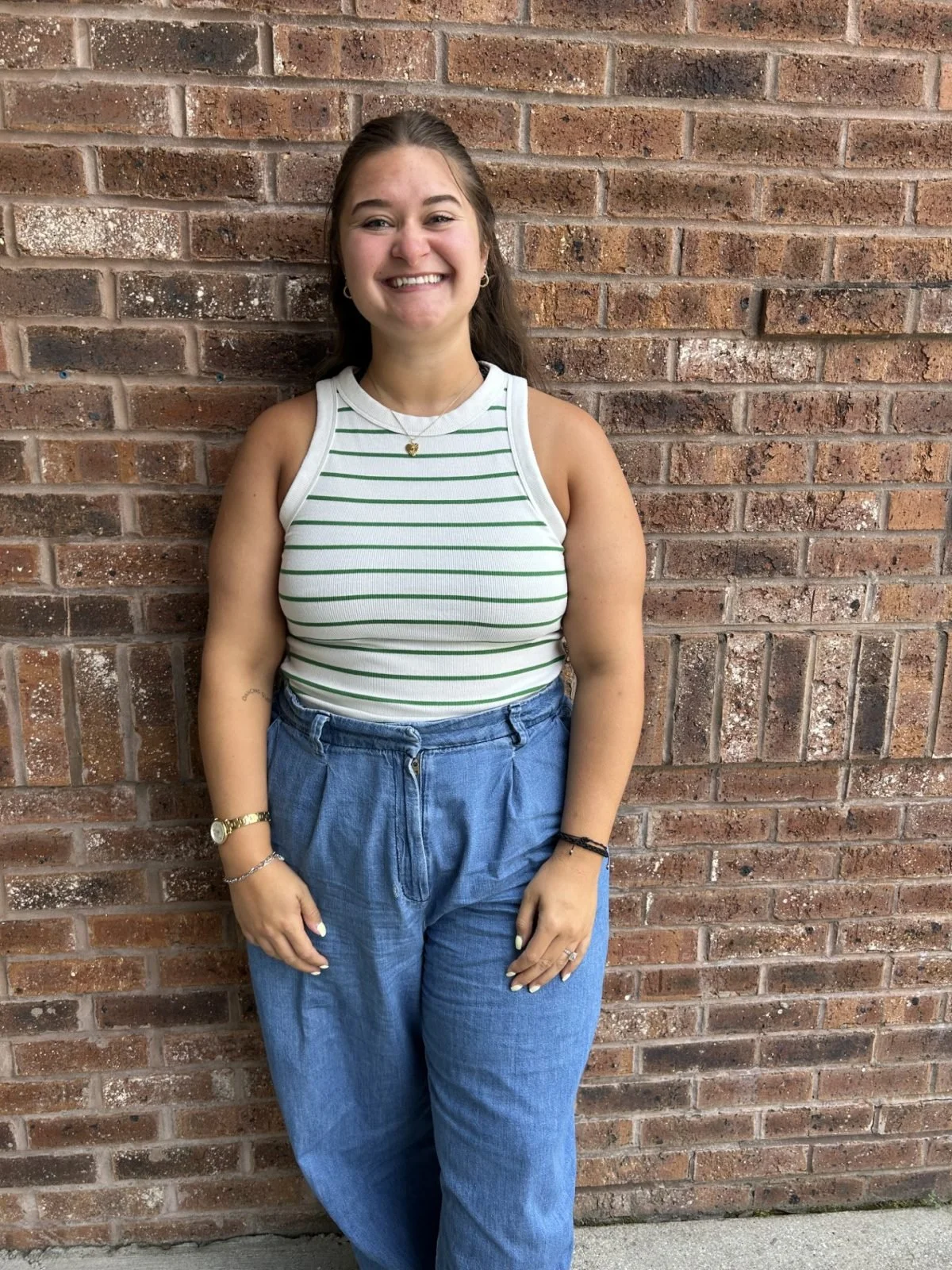 Young woman with long brown hair standing against a brick wall, smiling, wearing a sleeveless white and green striped top and blue jeans.