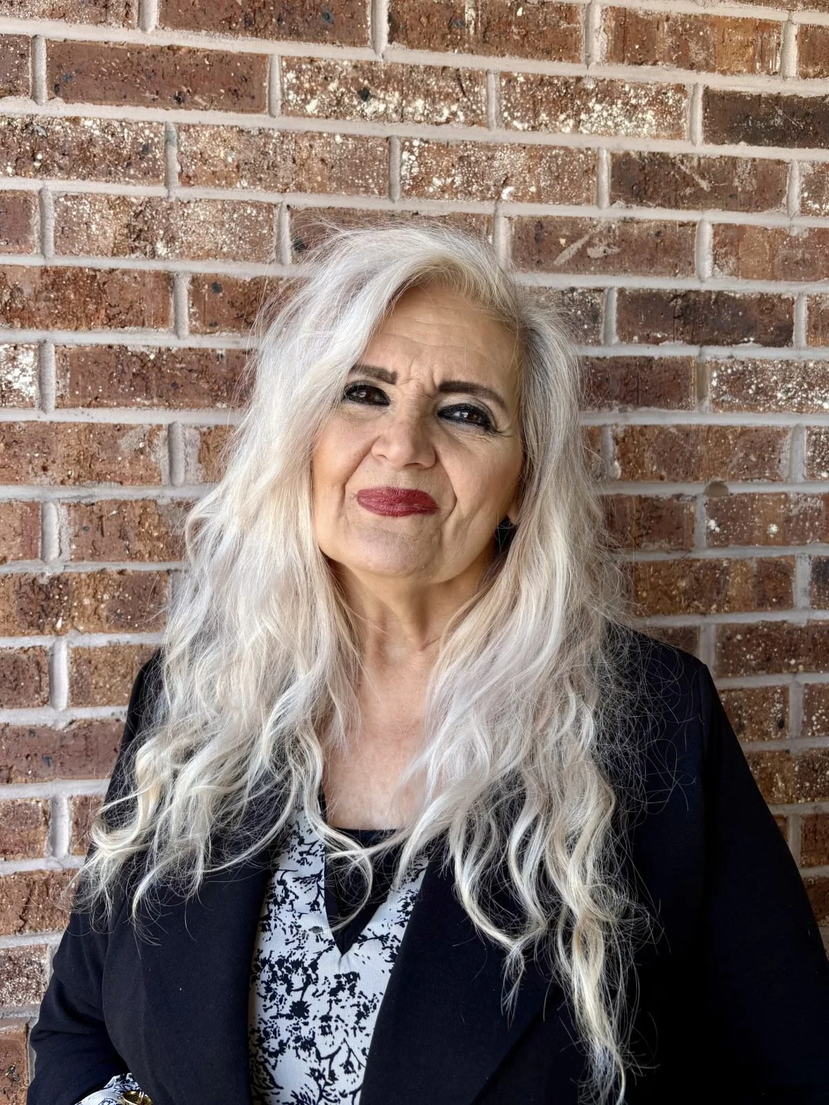 Portrait of an older woman with long, wavy gray hair, wearing a black blazer over a black and white patterned top, standing against a brick wall.