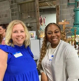 Two women standing indoors in a rustic setting, smiling at the camera. One woman has blonde hair and is wearing a bright blue top, while the other has dark hair styled in curls and is wearing a light-colored top and cardigan.