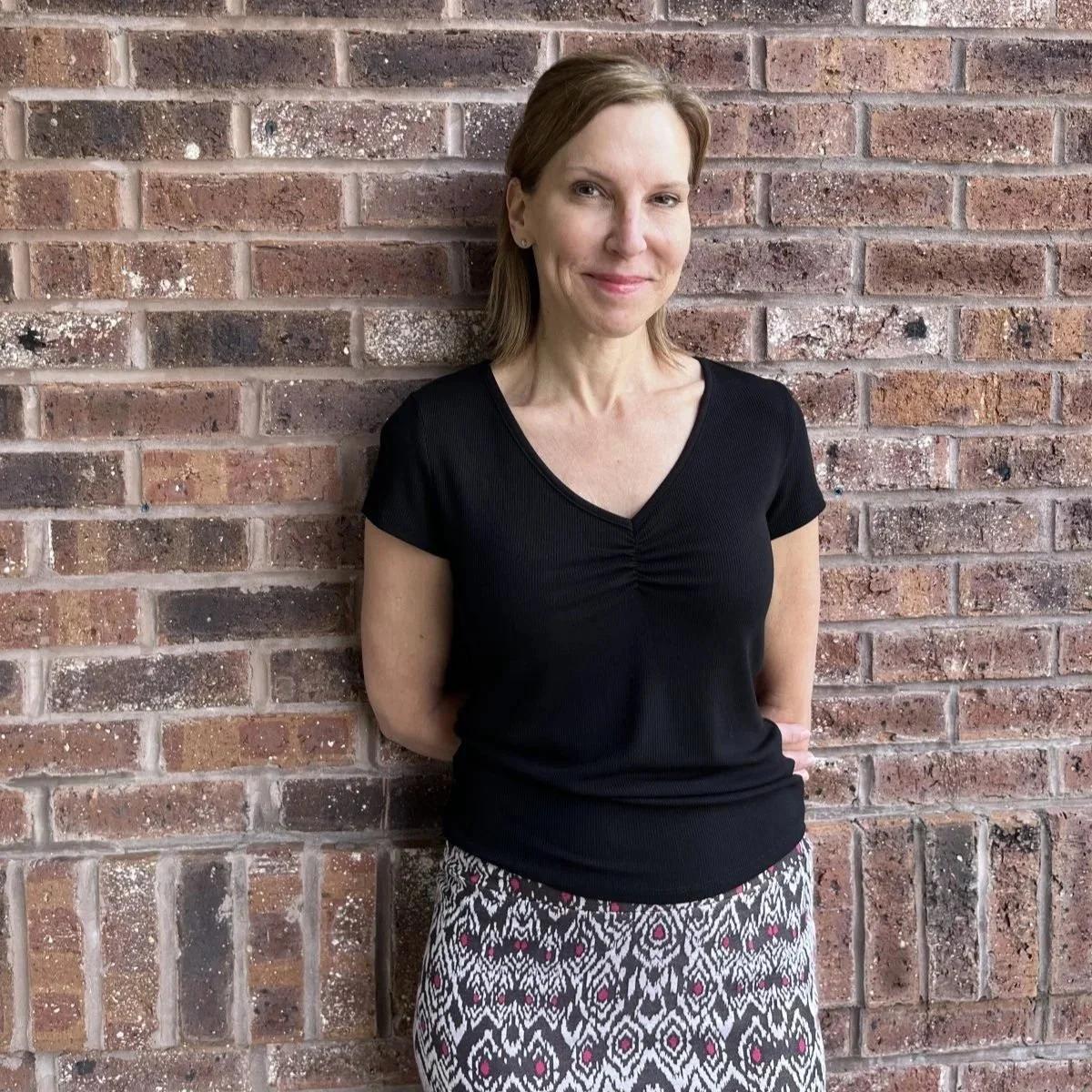 A woman with light skin and brown hair standing against a brick wall, wearing a black V-neck top and patterned skirt.