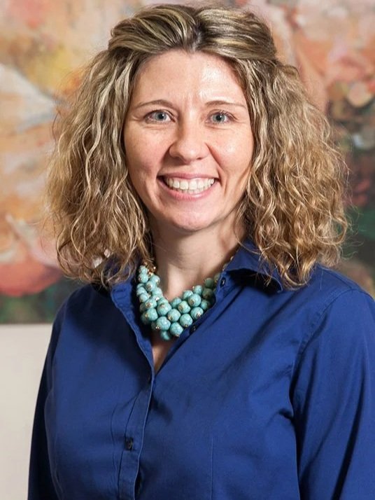 A woman with curly blonde hair wearing a blue button-up shirt and a turquoise bead necklace.