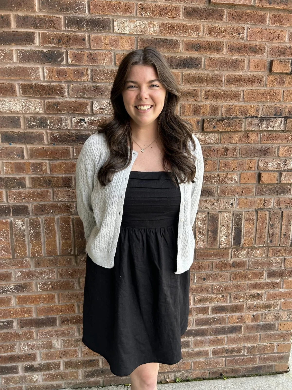 A young woman with long brown hair smiling, standing outdoors against an old brick wall, wearing a black dress, a white knitted cardigan, and a delicate necklace.
