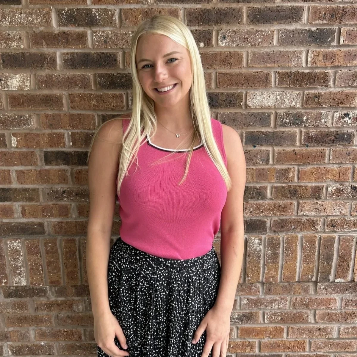 A young woman with long blonde hair, smiling, wearing a pink sleeveless top and a black skirt with white polka dots, standing in front of a brick wall.