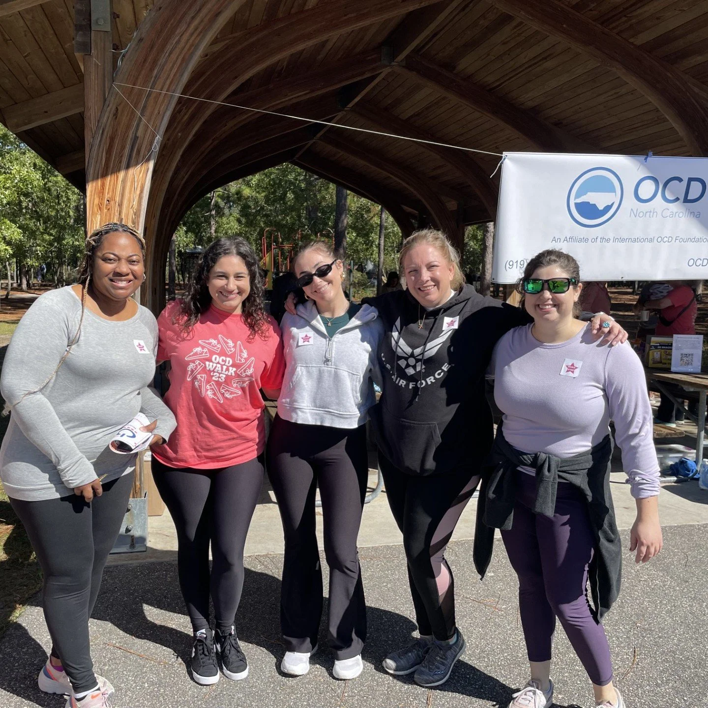 Group of five women smiling at an outdoor event, standing under a wooden shelter with a banner for North Carolina OCD support group behind them.