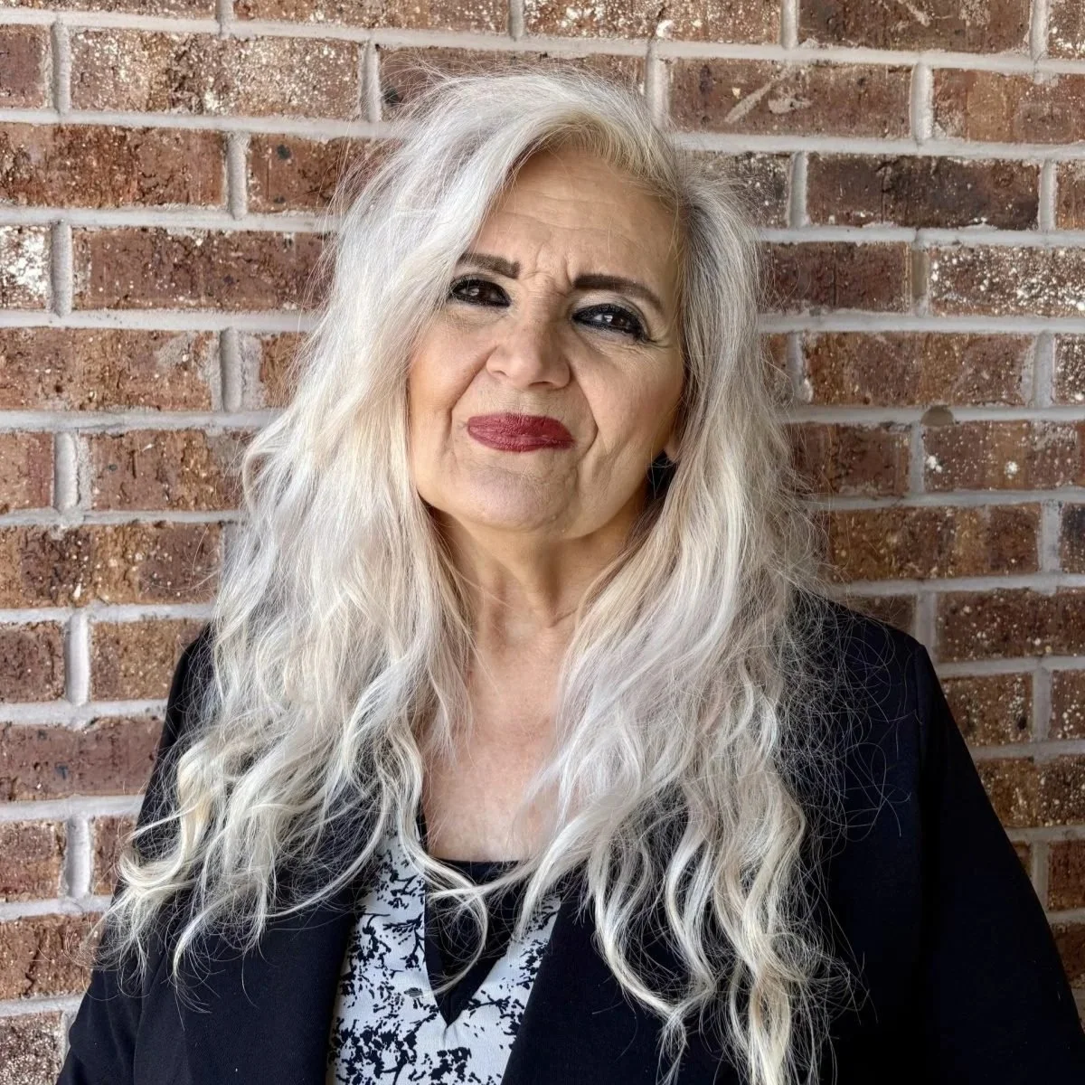 An older woman with long, wavy, blonde hair and dark makeup, standing against a brick wall.