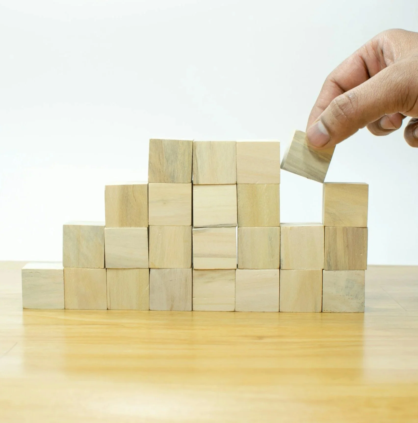 A hand removing a wooden block from a stack of small wooden cubes arranged in a stepped pattern on a wooden surface.