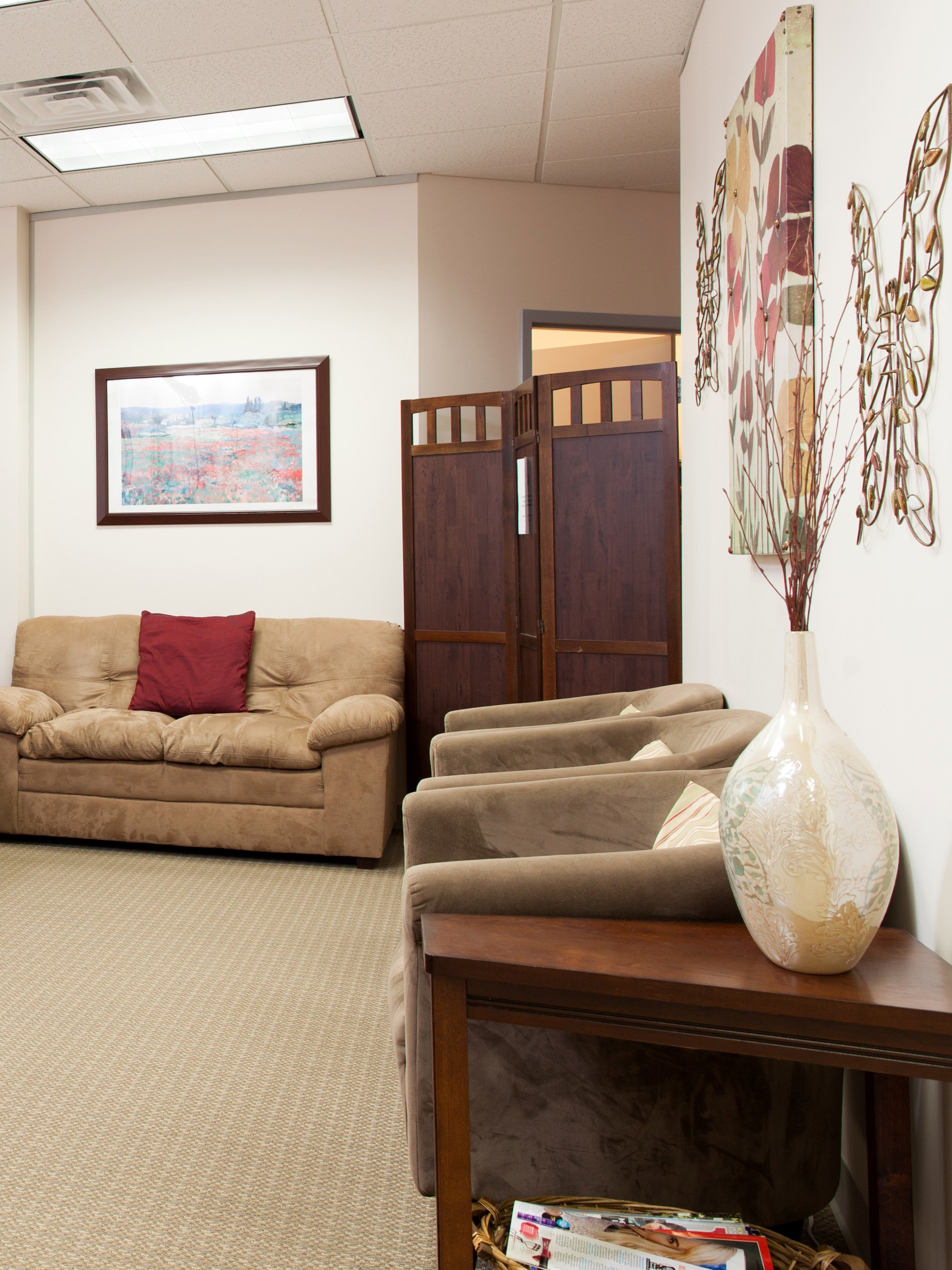 A cozy waiting room with beige sofa and chairs, a wooden room divider, artwork on the walls, and a large vase with decorative branches on a wooden side table.